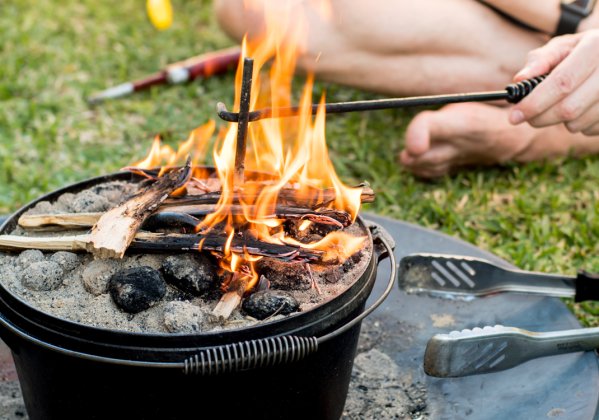 Dutch oven campfire cooking with flames and the legs and hand of a person holding a poker